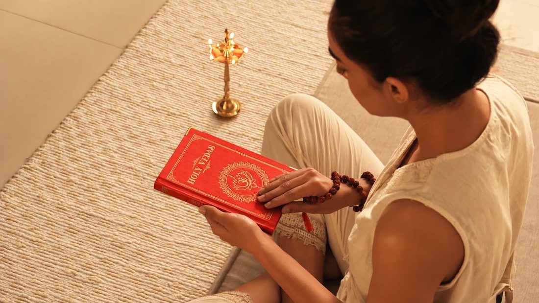 Donna seduta legge il libro Holy Vedas dalla copertina rossa accanto a una lampada ayurvedica in ottone accesa, per Art of Vedas